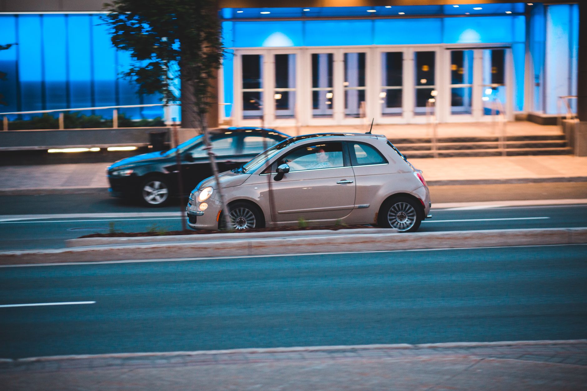 Fiat 500 jedzie nocą ulicą obok nowoczesnego, niebiesko podświetlonego budynku Fiat 500 jedzie nocą ulicą obok nowoczesnego, niebiesko podświetlonego budynku